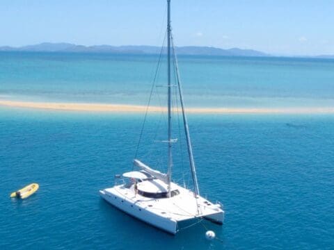 A white catamaran, Blizzard, is anchored in the clear blue waters of the Whitsundays near a small sandbar with a distant mountainous landscape. A yellow kayak floats nearby.