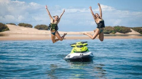 Two people in swimwear and life vests jump into the air next to a jet ski on a sunny day, bringing all the excitement one could expect from watersport safaris. In the background, a sandy shore and lush greenery complete the picturesque scene.