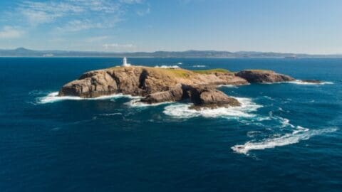 A small rocky island with a lighthouse sits in the blue ocean, surrounded by waves. The mainland is visible in the distant background under a clear sky, making it an ideal spot for an ecotourism tour.