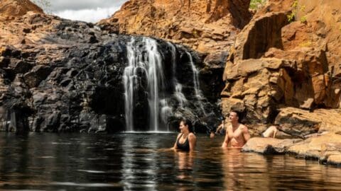 Two individuals enjoy swimming by a waterfall nestled among rocky cliffs, embarking on inspiring journeys amidst nature's serene beauty.