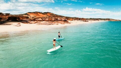 Two people paddle boarding on turquoise water near a sandy beach with rocky cliffs in the background under a partly cloudy sky, enjoying the serene beauty of an eco resort.