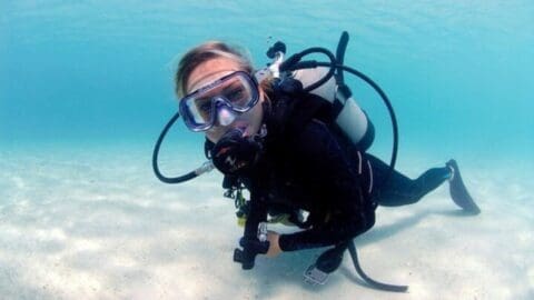 A person in certified scuba gear dives underwater in the clear, blue ocean, exploring a vibrant and sustainable marine environment.