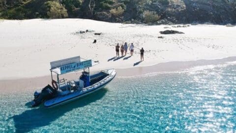 A small boat is docked on a clear, sandy beach. Several people stand on the shore near the boat, likely part of a guided tour. Trees grow on a hill in the background, offering a picturesque view for visitors enjoying sustainable accommodation nearby.