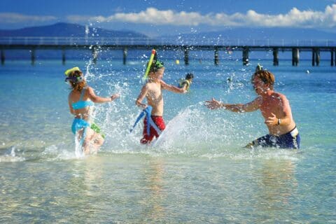 Three people, wearing snorkel gear, playfully splash water at each other in a shallow part of a seaside beach. A wooden pier extends into the water in the background, showcasing an ideal spot for ecotourism and nature exploration.