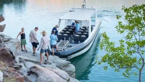 A group of people disembarks from a motorboat onto rocky terrain beside a body of water, with one person assisting others as they step off the boat. The serene atmosphere showcases the beauty of nature, creating a tranquil experience for everyone involved.