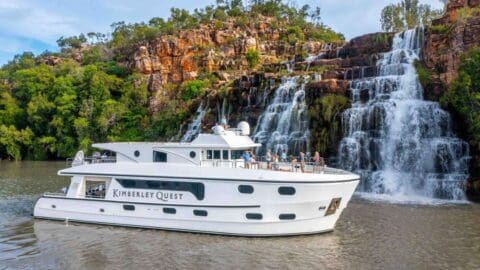 The Kimberley Quest, a white boat, is docked near a multi-tiered waterfall surrounded by lush greenery and rocky terrain, highlighting the beauty of nature and the benefits of ecotourism.