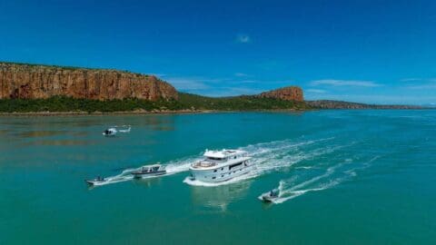 A large yacht cruises on a turquoise sea near a coastal cliff, accompanied by smaller boats and a helicopter flying under a clear blue sky, promoting sustainable ecotourism.