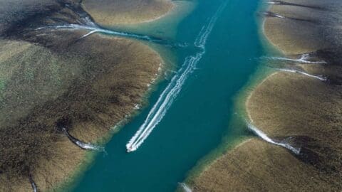 A boat navigates through clear blue water flanked by brown, circular land formations, leaving white wakes behind. This journey highlights ecotourism efforts, emphasizing sustainable practices and the natural beauty of the environment.