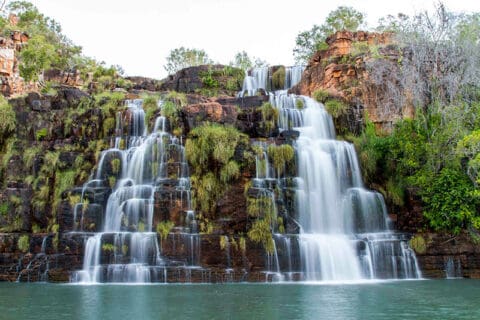 A multi-tiered waterfall cascading over a rocky cliff surrounded by lush greenery, with water flowing into a tranquil pool at the bottom—a perfect sight to encounter on an adventure with One Tide Charters.