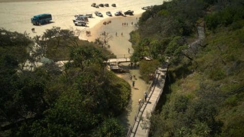 Aerial view of tourists exploring a sandy beach area near a small water body, with vehicles parked nearby and a wooden walkway leading into the trees, highlighting the beauty of nature.