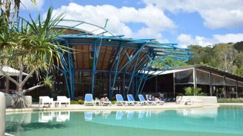 A resort building with unique blue steel architecture offers sustainable accommodation and a poolside area featuring lounge chairs under a partly cloudy sky.