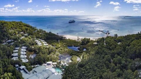 Aerial view of a coastal resort surrounded by lush greenery with a long pier extending into the ocean, and a cruise ship anchoring near the shore. This destination offers sustainable tours amidst nature under a partly cloudy sky.