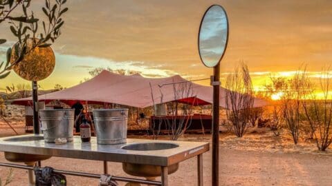 At sunset, the outdoor campsite, set up for World Expeditions, features a table with metal containers and bottles in the foreground and large tents neatly arranged in the background.