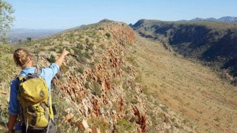 A person wearing a backpack, likely on one of their World Expeditions, points towards a distant view while standing on a rocky ridge with a vast desert landscape extending under a clear sky.