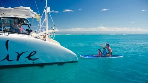 A person with two children on a paddleboard near a sailboat in clear blue water, embracing the beauty of nature on a sunny day.