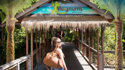 A woman with a large bag over her shoulder walks towards the entrance of Magnums Airlie Beach under a thatched roof sign, known for its commitment to ecotourism.