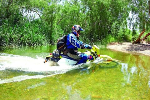 A motorcyclist wearing protective gear rides a yellow Suzuki dirt bike through a shallow, clear water stream surrounded by lush vegetation, showcasing an adventurous tour.