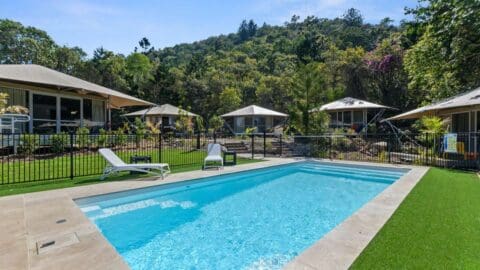 Outdoor swimming pool with lounge chairs, surrounded by a safety fence and lush greenery, featuring cozy cabins in the background and a forested hillside beyond. Perfect accommodation for an ecotourism getaway.