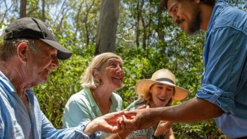 A group of people in casual attire are smiling and socializing outdoors on a sunny day, surrounded by greenery. One person is holding out their hand, and the others are looking at it with interest, possibly discussing ecotourism opportunities.