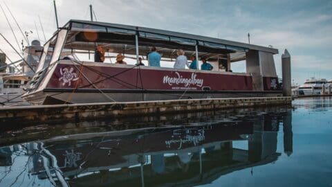 A maroon tour boat with people onboard is moored at a dock in a harbor, surrounded by other boats under a cloudy sky, promoting sustainable ecotourism.