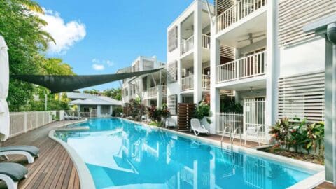 A blue pool with a sun screen surrounded by a wooden terrace and a modern white building.