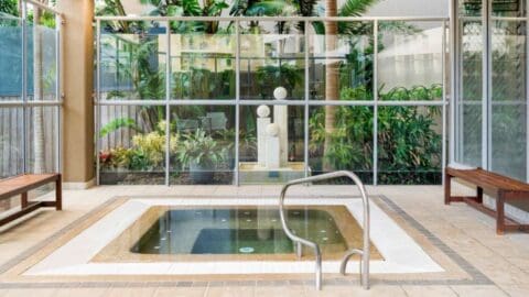 A calm jacuzzi surrounded by brown tile, large windows and a statue behind them.