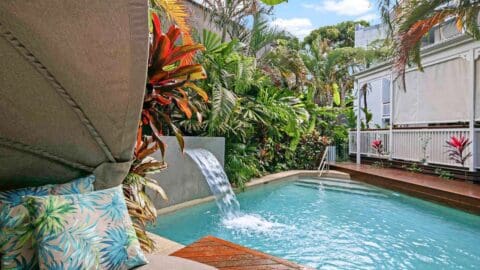 A fresh, crystal blue pool with a waterfall coming out of the wall surrounded by luscious plants.