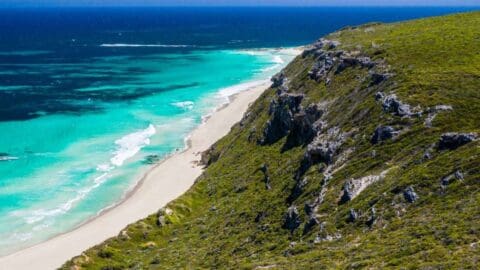 A coastline with vibrant turquoise waves hitting a sandy beach, bordered by rocky, sustainable cliffs under a clear blue sky.