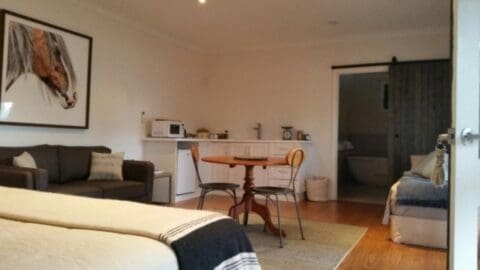 This cozy studio apartment features a comfortable bed, sofa, round table with chairs, kitchenette with appliances, and an open bathroom door. A framed horse painting hangs on the wall. Ideal for your next accommodation!