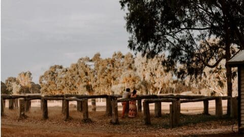 Two people standing near a wooden fence with trees in the background under a partly cloudy sky, embracing the beauty of nature.