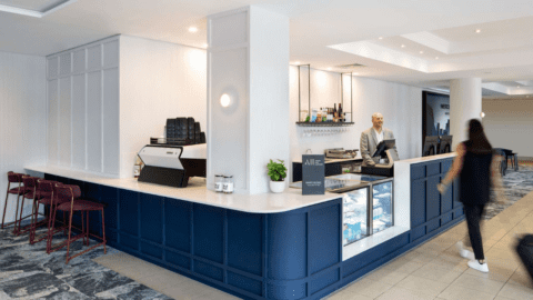 Hotel lobby café with a modern blue and white design at the Mercure in Garden City, Brisbane. A barista is behind the counter, and a customer is walking away. The café has seating, a coffee machine, and display cases with items.