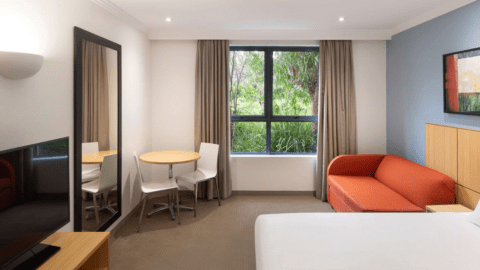A cozy Brisbane hotel room featuring a comfortable bed, a vibrant red sofa, and a small round table with two chairs. Enjoy entertainment on the wall-mounted TV next to a large mirror and relax by the window with curtains. Experience Mercure's touch in Garden City.