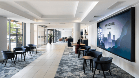 A modern hotel lobby with contemporary furnishings, including chairs and tables, welcomes guests at the Mercure Brisbane. A large sign with the hotel's name is mounted on the wall, capturing the essence of Garden City. The reception desk is visible in the background.