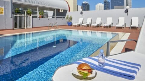 The rooftop pool area at Mercure Perth features lounge chairs and a table with a burger and water bottle atop a striped towel, offering a breathtaking cityscape view in the background.