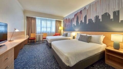 Experience the comfort of Mercure Perth with a hotel room featuring two double beds, modern decor, and stunning cityscape wall art. Enjoy a spacious desk by the large curtained window, two cozy chairs with a lamp in the corner, plush carpeted floors, and stylish bedside lamps for the perfect accommodation.