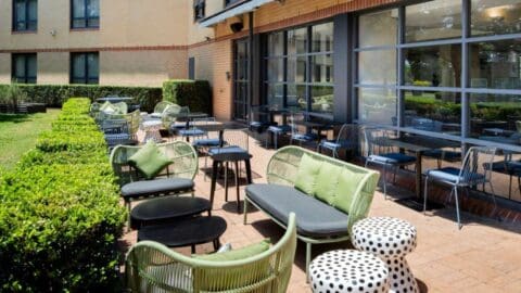 Outdoor patio area with green cushioned chairs, black tables, and polka-dot stools, adjacent to a building with large windows and surrounded by manicured hedges at Mercure Sydney Macquarie Park.
