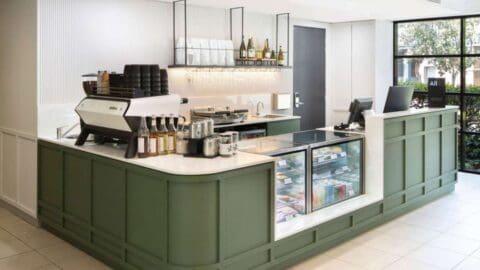 A modern coffee shop counter at Mercure Sydney features an espresso machine, various syrups, a display case with pastries, a cash register, and minimalist green and white cabinetry.