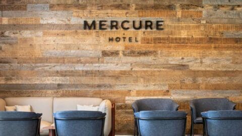 Seating area with gray chairs and a beige couch against a wood-paneled wall adorned with the "Mercure Hotel Sydney" sign.