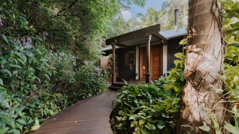 A wooden pathway leads to a small house with wooden doors, set amidst lush greenery and tropical plants. This charming accommodation offers an idyllic escape, perfect for an ecotourism adventure.