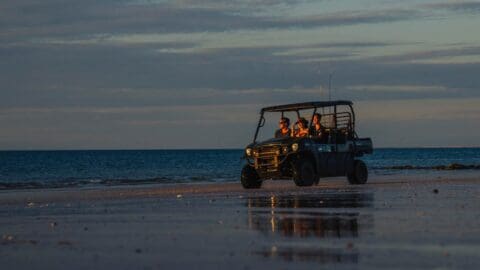 A group of people riding in an eco-friendly off-road vehicle along a beach at sunset, with the ocean in the background and reflections on wet sand.