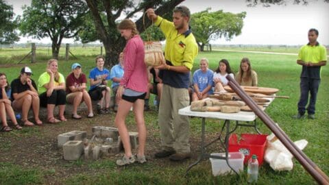 A group of people sits in a circle outdoors while a man in a yellow shirt demonstrates a tool to a woman in a pink shirt. Various equipment is placed on a table next to them, highlighting their tour of nature.