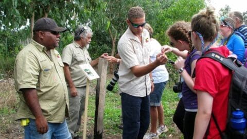 A group of people are gathered outdoors, observing and discussing something of interest. Some participants wear beige uniforms while others are in casual attire, including backpacks and cameras, immersing themselves in nature.