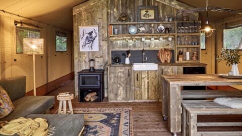 Rustic cabin interior with a kitchenette, wooden furniture, a stove, and hanging utensils. A couch and a coffee table are in the foreground. Warm, cozy ambiance that connects you to nature.