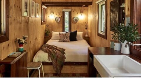 A cozy wooden cabin bedroom with a made bed, a small sink area, a side table with decorations, and windows providing natural light and views of nature.