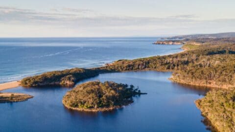 Aerial view of a coastal landscape featuring a calm bay, a small island, and a forested shoreline with distant hills under a clear sky, perfect for an accommodation in nature or an engaging tour.