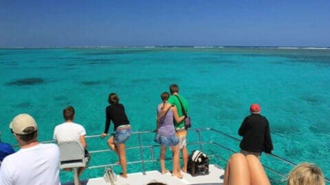 A group of people on a boat's deck looks out over a vast expanse of clear turquoise water during a sustainable tour on a sunny day.