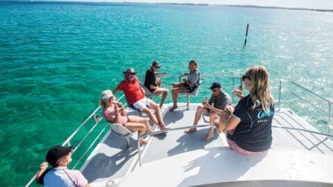 A group of people enjoy the beauty of nature, sitting and standing on the deck of a boat in the middle of a clear, turquoise sea during a sunny day tour.