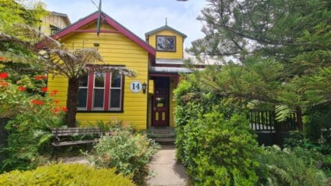 A yellow house with a red front door, marked with the number 14. The lush yard features greenery, bushes, and tall trees. A bench is placed on the left side near the front entrance. This charming accommodation promotes sustainable living practices.