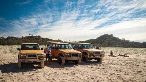 Three beige off-road vehicles are parked on a sandy beach, part of a sustainable tour. People stand near the vehicles, while two individuals lie on the sand. Hills and trees serve as a picturesque backdrop under a blue sky.