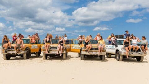A group of people in swimwear sits on and around parked trucks at a sandy beach under a partly cloudy sky, as part of their coastal tour.